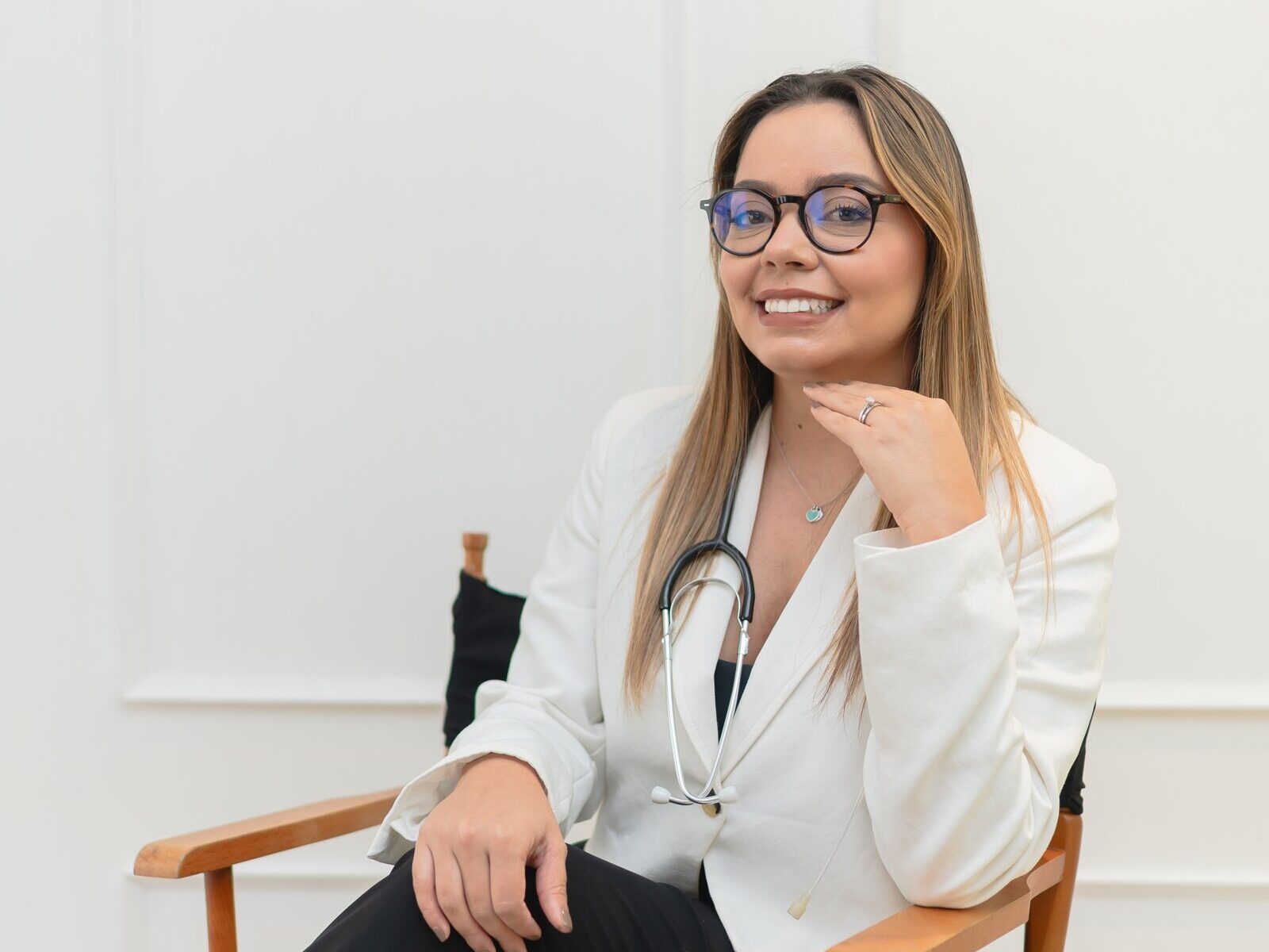 Deylin Mena Vega, Máster en Audiología, sentada y sonriendo, con saco blanco y estetoscopio